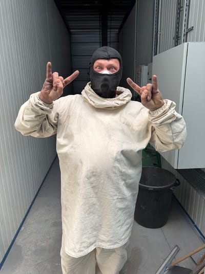 A person, wearing a tan protective suit and a black balaclava with a mask, stands in what appears to be a storage unit or industrial space in Mol, Belgium. The person is making a "rock on" hand gesture with both hands, with their thumbs extended and index and pinky fingers raised. Their eyes are visible through the mask and appear wide open, looking directly at the camera. The suit is loose-fitting and appears to be made of a textured fabric. The background consists of light-colored corrugated metal walls, a concrete floor, and some industrial equipment, including a large black plastic bin and what looks like electrical panels. The lighting is somewhat dim, suggesting an indoor environment. The overall impression is of a person in protective gear making a defiant or celebratory gesture in a utilitarian setting.