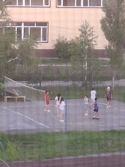The image shows a group of young people on an outdoor sports court, possibly a basketball court due to the markings on the ground. There are at least six individuals visible. Some are standing, while others appear to be in the process of or have just completed an activity, possibly a race or a drill, as some are lying down.
The individuals are dressed in casual sportswear. One person is wearing a dark purple shirt and light shorts, another a white t-shirt and blue shorts, and another a white t-shirt and black shorts. There is also a person in a red t-shirt lying on the ground, and another in a white top. One person is wearing a light-colored hat.
The background includes a building with several windows, which appears to be an educational institution or dormitory, and a grassy area. The court is marked with white lines. The lighting suggests it is daytime, with no strong shadows indicating midday. The overall atmosphere appears to be one of casual activity, perhaps a sports practice or a break between classes. There is no visible text in the image. The location is Jitiqara, Kazakhstan.