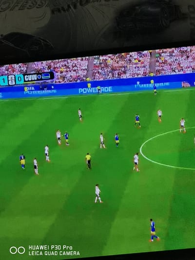 The image displays a broadcast of a soccer match, as evidenced by the green field, players in uniform, and a scoreboard at the top. The scoreboard shows the score as "1 0 CUWO", with a flag to the right of "CUWO" that appears to be the flag of Rwanda. A banner with the FIFA logo and "POWERADE" is visible along the edge of the field. The audience is in the stands behind the banner. The scene is not specifically located in the city of Bad Oeynhausen, Germany; rather, it is a generic soccer match broadcast. There are no elements in the image that would suggest a specific location within Bad Oeynhausen. The bottom of the image shows a watermark reading "HUAWEI P30 Pro LEICA QUAD CAMERA".