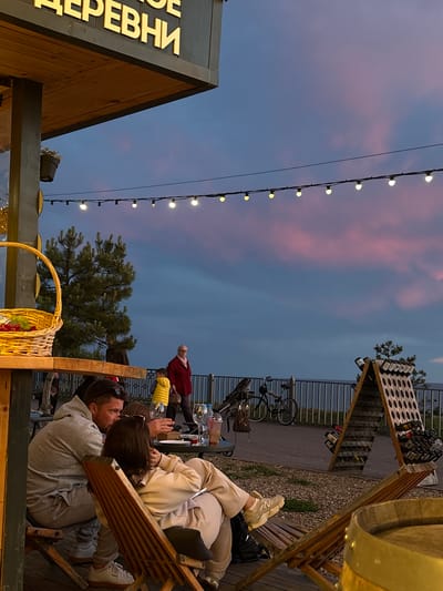 This image captures an outdoor evening scene at a cafe or winery in Akhtanizovskaya, Russia, during twilight. The sky exhibits soft blue and pink gradients, indicative of sunset or dusk, complemented by illuminated string lights overhead.

In the mid-ground, a man and a woman are seated closely on wooden chairs, facing each other in what appears to be an intimate or social interaction. The woman's legs are propped up on a wooden bench. Both are dressed casually. Other patrons are visible in the background, including a person in a red top and another in yellow, seated at tables.

The setting is an outdoor patio or terrace, furnished with wooden seating. Notable objects include a woven basket brimming with fruit (grapes and apples), various glasses on tables, and wooden racks designed to hold bottles, suggesting a wine-related establishment. A large, golden barrel-like structure is partially visible on the far right. Bicycles are parked near a metal fence in the background, and evergreen trees punctuate the scene.

A wooden structure on the left features golden Russian text. The visible portion reads "ОЕ ДЕРЕВНИ," likely part of a larger sign such as "ВИННОЕ ДЕРЕВНИ" (Wine Village), which aligns with the context of bottle displays and barrels. The overall ambiance is relaxed and social.