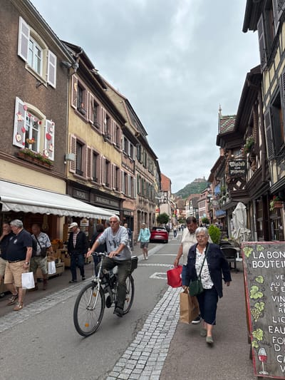 The image depicts a street scene in Ribeauvillé, France, a town known for its medieval architecture. In the foreground, a man is riding a bicycle down a cobblestone street. He is wearing a grey long-sleeved shirt and dark pants. To his right, an elderly woman walks towards the viewer, carrying a brown shopping bag and a green purse. She is wearing a floral shirt and dark pants. Behind them, a group of people are walking and browsing in front of shops. The buildings lining the street are characteristic of Alsatian architecture, with timber framing and colorful shutters. The sky is overcast, suggesting a cloudy day. In the background, a castle perched on a hill is visible, a common landmark in the Ribeauvillé area. The street is lined with shops selling various goods, including baked goods and souvenirs, as indicated by signage like "VOGEL BOULANGERIE PATISSERIE" and a chalkboard sign on the right advertising wine and food. The overall atmosphere appears to be that of a bustling tourist town.