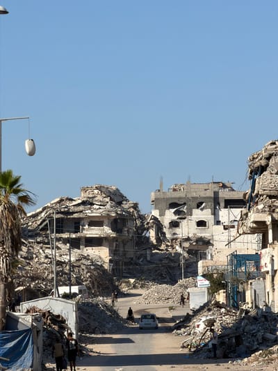 In Jabalia, Palestinian Territories, a street scene depicts significant destruction. Buildings are heavily damaged, with rubble and debris piled high on either side of a paved road. The sky is clear and blue.
On the left side of the image, a street lamp with a white, elongated fixture hangs from a pole. A palm tree with sparse fronds stands in front of partially collapsed structures. One building shows a partially intact concrete facade with numerous pockmarks, suggesting shrapnel damage. Arched windows are visible, some boarded up.
In the center of the scene, a white van is parked on the road, and a white car drives away from the viewer. Several individuals are walking on the street and near the rubble. Two young males are walking towards the viewer in the foreground, carrying bags. Another individual, a man, is standing by a pile of debris, seemingly observing or taking pictures.
On the right, more damaged buildings are visible, with exposed rebar and concrete. A blue metal structure, possibly part of scaffolding or a temporary shelter, is present. A white sign with Arabic text is attached to one of the damaged buildings. The overall impression is one of widespread destruction and the ongoing efforts of people navigating the aftermath.