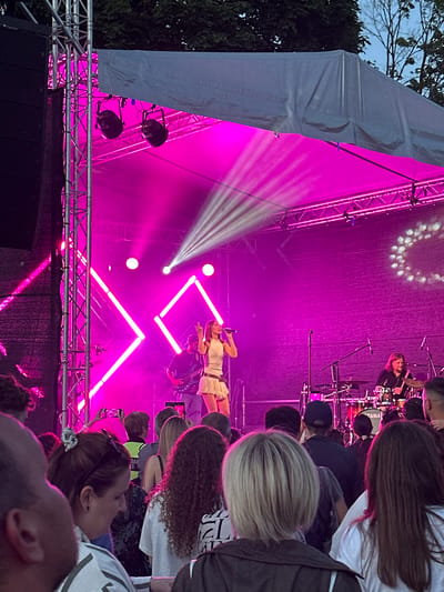 The image captures a live music performance during an outdoor event, possibly a festival, at dusk in Panevėžys, Lithuania. The stage is bathed in vibrant pink lighting, with bright white spotlights illuminating the performers. A female singer, dressed in a white outfit, stands center stage with a microphone, seemingly in the midst of a song. Behind her, a drummer is visible playing a drum kit. The stage features geometric lighting structures, adding a modern aesthetic to the performance.
In the foreground and surrounding the stage, a crowd of spectators is gathered, their faces partially obscured by the low angle of the shot and the dim lighting. Many are looking towards the stage, engrossed in the performance. Some are holding up their phones to capture the moment, suggesting it's a significant performance for them. The presence of trees and a cloudy sky visible in the background indicates an outdoor setting. The overall atmosphere appears energetic and engaging, with the bright lights and the crowd's attention creating a sense of shared enjoyment. The time of day is likely early evening, given the sky's color and the artificial lighting taking prominence.