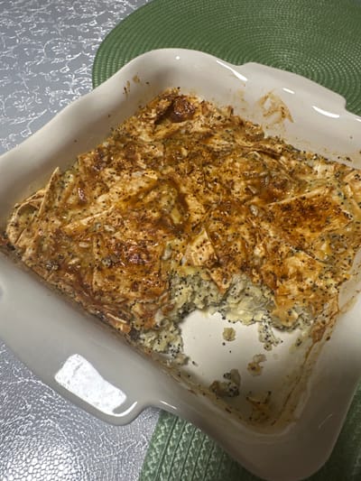 A partially eaten poppy seed pastry is in a white, square baking dish with rounded corners. The dish is sitting on a silver, textured surface. A green, round woven placemat is partially visible on the left side of the dish, and a green placemat is on the right side. The pastry has a golden-brown, flaky crust with poppy seeds scattered on top. The filling appears to be a light-colored mixture with poppy seeds. A portion of the pastry is missing from the top right. The baking dish is slightly stained on the interior. The location is not discernible in the image.