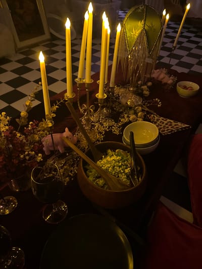 The image depicts an intimate, dimly lit indoor dining scene, viewed from an overhead, slightly angled perspective. A long table, covered with a dark red tablecloth, is richly adorned with decorative and dining elements.
A prominent multi-arm candelabra, holding several electronic candles, emits a warm yellow glow, illuminating the immediate area. To its right, a wooden salad bowl filled with green salad and crumbled white cheese contains two wooden salad servers. Nearby, a small white bowl holds what appears to be red berries or fruit. A black dinner plate is positioned to the right of the salad bowl.
Scattered across the table are dried floral arrangements featuring delicate white baby's breath and reddish-brown foliage. A small, light pink pig-shaped figurine is nestled among these decorations. Several wine glasses, some containing a dark liquid, are also visible. Other decorative elements include a green, ornate lampshade or a similar item with beaded fringes, and a piece of leopard-print fabric or napkin.
The background reveals a distinctive black and white checkered floor and a portion of a red cushioned seat or sofa, suggesting a comfortable and stylish dining area. A framed picture is partially visible on the wall in the lower left. The low, ambient lighting, predominantly from the candles, indicates an evening setting, creating a cozy, festive, or romantic atmosphere for a meal or gathering. No people or visible text are present in the image.