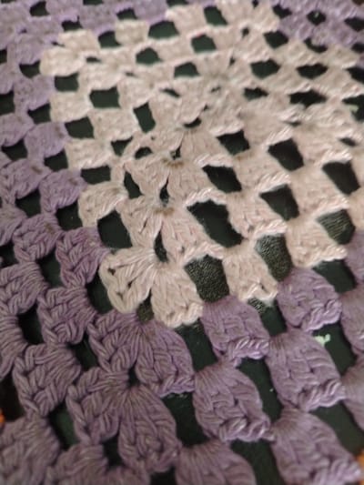 The image shows a close-up, blurred view of a crocheted textile. The textile features a repeating pattern of open squares formed by loops of yarn. The colors are primarily muted purple and pale pink, with dark spaces visible between the crocheted sections. The lighting is soft, and the focus is not sharp, creating a textured and slightly abstract appearance. There is no discernible information within the image to connect it to Stepnoe, Russia, or any specific location.