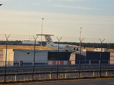 The image depicts an airport scene at the city of Kelsterbach, Germany, likely Frankfurt Airport, given its proximity and common aircraft types. In the foreground, a chain-link fence with barbed wire runs horizontally across the frame, partially obscuring the view. Behind the fence, several portable buildings or shipping containers are visible. The largest building is a light-colored, rectangular structure with a door and a small window. Adjacent to it are darker colored containers, one of which is navy blue.

In the midground, an airplane is parked. It is a narrow-body jet with a distinctive snowflake logo on its tail. The text "D-NICE" and "NICE Training" are visible on the fuselage. The aircraft appears to be stationary. Two red and white striped poles, likely navigational markers or lighting, are positioned near the aircraft. The ground surrounding the containers and aircraft is covered in dry grass and some patches of asphalt.

In the background, more buildings and trees can be seen, suggesting the expanse of the airport or surrounding area. The sky is mostly clear with a few wispy clouds, illuminated by the warm light of what appears to be late afternoon or early evening sun. The overall impression is one of an operational but quiet section of an airport. There are no people or animals clearly visible in the scene.