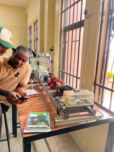 The image shows two men looking at a smartphone on a table indoors. The man in the foreground wears a beige polo shirt. A man in a green and white baseball cap is partially visible next to him. On the table are books and a red motor with a clear plastic electrical housing, part of an apparatus of some kind. The location could be Afaka, Nigeria, based on the prompt. Through the windows, a building with a red tile roof and metal security bars is visible. The interior walls are painted beige.