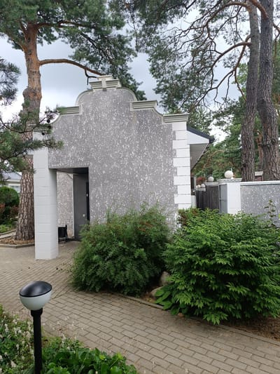 The image depicts an overcast outdoor scene in a residential area, likely in Старобінскі сельскі Савет, Belarus, judging by the architecture and vegetation. In the foreground, a modern pathway made of grey rectangular pavers leads towards a building entrance. A black and white outdoor light fixture on a pole stands to the left of the pathway, surrounded by low-lying green plants and some dried white flowers.
The main subject is a building with a gray, textured facade, featuring a prominent white architectural trim around its roofline and entryway. The entrance is an open doorway, dark inside, with a light switch visible on the right-hand interior wall. White, square-shaped columns flank the entrance. To the right of the entrance, a dense green bush occupies the mid-ground.
Behind the building, a grey fence with decorative white posts topped with spherical lights is visible. Beyond the fence, more greenery and the roofline of another house can be seen. Tall pine trees with rough bark dominate the background, their branches extending over the scene. The ground is covered with pavers and some scattered pine needles. No people are present in the image.