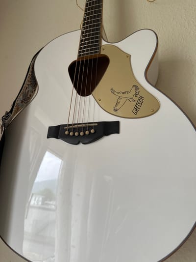 This is a close-up, eye-level shot of a white Gretsch acoustic guitar. The guitar is positioned diagonally in the frame, with its body taking up most of the image. The pickguard, a gold-colored teardrop shape, is prominently displayed on the guitar's body, featuring the Gretsch logo and an illustration of two birds in flight. The guitar's bridge, made of black material, holds six strings that extend up the neck, which is partially visible on the left side of the frame. The guitar's body is smooth and highly reflective, showing a distorted reflection of what appears to be a room with furniture. A strap, adorned with a floral and patterned design, is attached to the guitar at the bottom. The background is a textured, off-white wall, suggesting an indoor setting. The lighting is neutral, with no strong shadows, and the overall impression is of a well-lit interior space. There is no specific activity or event taking place, as the image focuses solely on the object. The text "GREISCH" is visible on the pickguard, printed in a stylized font. The location context of Bad Blankenburg, Germany is not directly evident from the visual information in the image itself, but is provided as external information.
