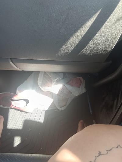 The image shows the interior of a vehicle, likely a car, with a person's leg and foot visible in the foreground. The flooring of the vehicle appears to be a dark-colored rubber mat. A white plastic bag, possibly containing items, is situated on the floor beneath the dashboard, partially illuminated by sunlight streaming through the vehicle's windows. A red and white shoe is also visible on the floor. The overall lighting suggests it is daytime. The scene is captured from a low-angle perspective, looking into the footwell of the vehicle. There are no visible landmarks or contextual clues that would allow for the identification of a specific location within Rock Springs, United States of America.
