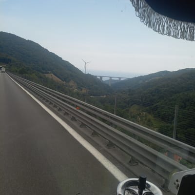 The image captures a view from a moving vehicle on a highway in Mele, Italy, likely on the A10 Autostrada. The road ahead is paved and has a white lane marker. To the right, a metal guardrail runs alongside the road, separating it from a steep, tree-covered hillside. Beyond the hillside, a large concrete bridge spans a valley, and further in the distance, a wind turbine is visible against a hazy sky. The sky itself is bright and overcast. The vehicle appears to be a bus or similar large passenger vehicle, as suggested by the fringe visible at the top of the image, which is likely part of the vehicle's interior upholstery. A small, chrome-plated object, possibly an ashtray or a part of the vehicle's dashboard, is visible in the lower right corner of the image.