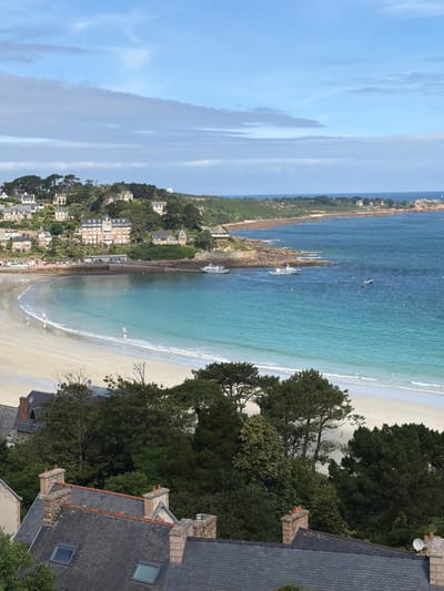 The image depicts a scenic coastal view of La Clarté, France. The scene is dominated by a tranquil turquoise sea meeting a sandy beach, with lush green trees and houses dotting the hillside in the background. The sky is a clear blue with wispy white clouds, suggesting a pleasant, sunny day. Several boats are visible in the water, some anchored and others appearing to be in motion. Small figures of people can be seen on the beach, enjoying the seaside. The foreground features the rooftops of houses, with chimneys and dormer windows, hinting at a residential area overlooking the coast. The overall impression is one of a peaceful, idyllic vacation spot. The time of day appears to be late morning or early afternoon, given the brightness and angle of the sun.