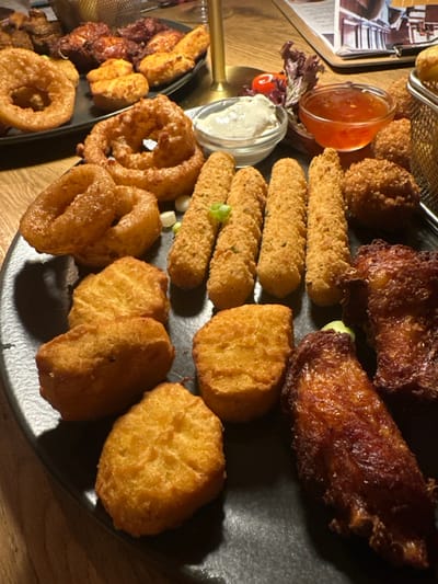 The scene depicts a close-up view of multiple fried food items arranged on black serving platters, consistent with a restaurant environment in Weißenburg in Bayern, Germany. In the foreground, a large, irregularly shaped black platter displays a variety of items. These include approximately seven golden-brown onion rings of varying sizes, five elongated golden-brown breaded mozzarella sticks garnished with chopped green onions, approximately seven golden-brown chicken nuggets of irregular shapes, and two dark reddish-brown fried chicken wing segments. Two small clear glass bowls are present on this platter; one contains a white creamy dipping sauce, and the other holds a reddish-orange sweet chili-style sauce. A second, partially visible black platter in the upper left background shows additional fried items, including more chicken wings and small, rounder fried pieces. A golden-colored stand is positioned between the platters. In the upper right background, a blurred menu or leaflet with text and images is visible. All items are situated on a light-colored wooden table surface under indoor lighting. No individuals are present in the frame.