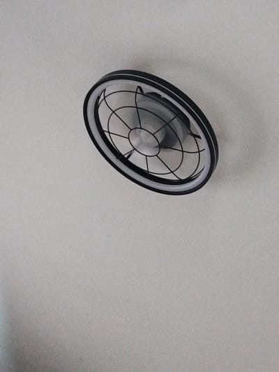 This is an eye-level, slightly angled medium shot of a modern ceiling fan. The fan is circular with a black outer rim and a white inner ring that appears to be a light fixture. The central part of the fan has a black grill that forms a starburst pattern. The blades of the fan are visible within the grill, and they are dark in color, possibly black or dark gray. The background is a plain, textured white ceiling, suggesting an indoor setting. The lighting is soft and diffused, with no harsh shadows, indicating it could be daytime with indirect natural light or the artificial light from the fan's fixture itself. There is no indication of people, specific weather conditions, or time of day beyond what can be inferred from the lighting. The setting appears to be a room in a home or an office. No text is visible in the image.
