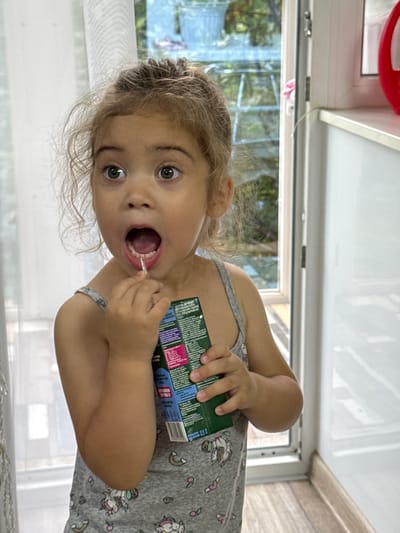 A young girl with blonde curly hair and wide, surprised eyes is shown in a close-up, waist-up shot. She is wearing a gray tank top with a unicorn and rainbow pattern. Her mouth is open as she holds a small, white object, possibly a toothpick, near her teeth. In her right hand, she clutches a small, green cardboard box with text in various colors. The background is softly blurred, showing a white curtain on the left and a window on the right, through which greenery and a railing can be seen. The flooring appears to be light brown wood. The lighting suggests an indoor setting with natural light coming from the window. The scene is likely captured in a home environment. No specific location in Ishimbay, Russia, can be determined from the visual information alone.