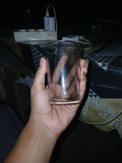 In Teungkudibandapirak, Indonesia, a person's left hand holds a clear glass. The glass is partially filled with a liquid, visible as a darker area at the bottom and sides, with some condensation forming on the exterior. The hand is positioned with the palm facing inwards, supporting the glass from below. The person's fingernails are long and appear to be unpainted. In the background, a power strip with multiple outlets and a white USB charger plugged in are visible. The surface beneath the power strip and glass is covered with a patterned cloth. The lighting is dim, suggesting an indoor setting at night.