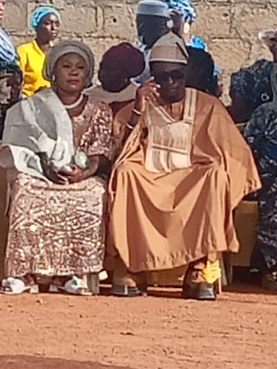 This image depicts an outdoor gathering in Afaka, Nigeria. In the immediate foreground, two adults are seated side-by-side on low chairs or stools upon reddish-brown unpaved earth.

The individual on the left is a female, dressed in a light grey headwrap (gele), a long-sleeved dress adorned with shiny, light-brown or bronze-colored sequins or embellishments, and a light-colored shawl draped over her left shoulder and lap. She wears a white beaded necklace and white sandals with a green detail. Her hands are clasped in her lap, holding a small, light-green object. Her face is visible, looking slightly towards her right.

The individual on the right is a male, wearing a tan or light brown traditional agbada or kaftan, which features a lighter, possibly embroidered, vertical panel on the chest. He wears a matching skullcap (fila) and dark sunglasses. His right hand is raised, with fingers near his ear or the side of his face. He wears dark sandals with a yellow or gold accent on the strap of his right foot.

In the mid-ground, behind the two seated figures, several other individuals are partially visible. To the left, a woman in a bright yellow top and a light blue headwrap is visible. Other figures are less distinct, dressed in various traditional attire. The background consists of a light-colored, textured stone or concrete wall. The scene is illuminated by natural daylight.