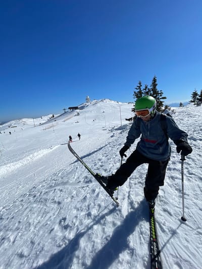 A male skier is captured in a low-perspective photograph on a snow-covered mountain slope in Jahorina, Bosnia and Herzegovina, under a clear, bright blue sky. The individual, positioned slightly right of center, wears a light green ski helmet with white-framed goggles featuring orange lenses, a dark grey or blue quarter-zip top with a red logo on the chest, black ski pants, and black gloves, with a dark backpack visible. He stands on black and green "Elan" skis, with his right ski lifted at the tail while his left ski remains flat on the snow. He holds two silver ski poles and displays an open-mouthed expression, revealing teeth. In the background, the snow-covered slope ascends towards a distant peak, where a prominent white, dome-shaped building and an adjacent structure are visible, consistent with a weather station or observatory. Multiple ski lift towers and cables, including a multi-seat chairlift station, are present on the ascending slope. Several other individuals are discernible skiing or walking further up the mountain. Sparse conifer trees and brown wooden ski fences line parts of the slope. Long shadows are cast across the snow from the skier and background features.
