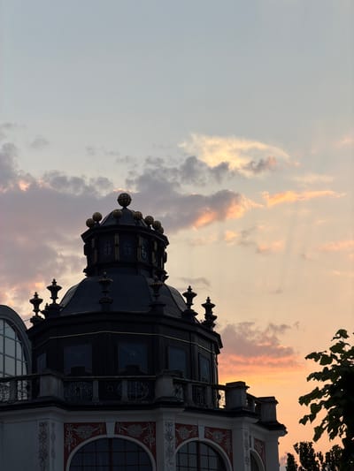 The image captures a detailed view of the dome of the Krzywy Domek (Crooked House) in Sopot, Poland, during sunset. The sky is filled with soft clouds illuminated by the warm hues of the setting sun, casting a gentle light on the architectural details. The dome itself is dark, possibly black or dark blue, with ornate golden accents and decorative spherical elements on its top. Below the dome, a balcony with intricate railings is visible, and further down, the facade of the building is adorned with red and white decorative patterns, framing arched windows. To the right, a cluster of trees with dark green leaves is silhouetted against the colorful sky, with the tops of other trees visible in the distance. The scene is static, with no people or actions depicted. The focus is on the architectural grandeur of the building juxtaposed with the serene beauty of the sunset sky.