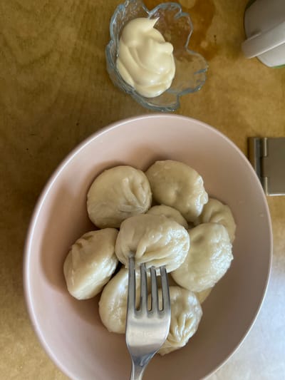 In Osinniki, Russia, a bowl of manti, a type of dumpling, is served on a table. A fork is piercing one of the manti, suggesting they are ready to be eaten. Adjacent to the bowl of manti, a small glass dish contains a dollop of white sauce, likely sour cream or mayonnaise, commonly served as a condiment with manti. The table surface appears to be made of wood, with a warm, brown hue. In the background, a portion of a white cup is visible, possibly part of a tea or coffee set. The lighting suggests an indoor setting, likely a dining area.