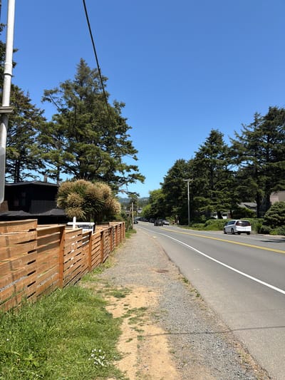 A clear, bright day is depicted in this photograph of a residential street in Cannon Beach, Oregon, United States of America. The road, which runs parallel to a grassy shoulder and a wooden fence, is flanked by tall evergreen trees on both sides. On the left side of the road, a wooden fence encloses a property with a modern, dark-colored building. A white sign with some text is visible behind the fence. On the right side of the road, several cars are parked and driving. A white minivan is in the foreground, and a white sedan is further down the road. The sky is a vibrant blue with no clouds.