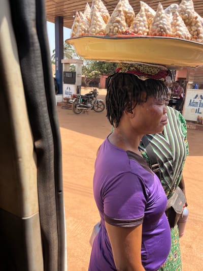 The scene depicts an outdoor street or marketplace environment in Otukpa, Nigeria. The foreground is dominated by a dark-skinned woman with dreadlocks, positioned in profile facing right. She is balancing a large, flat, yellow tray on her head, which holds approximately a dozen conical, clear plastic bags filled with light-colored nuts, likely groundnuts. A patterned headwrap is visible beneath the tray. She wears a purple short-sleeved top with dark trim and exhibits a closed-eye or squinting expression, with a noticeable mark on her right cheekbone.
Behind the primary subject, a second dark-skinned woman is partially visible, wearing a green, white, and black striped top and a patterned lower garment, with a dark bag slung over her shoulder. Further back and to the right, a third individual, a man in a red and white striped shirt, is faintly visible near a blue and white structure.
The background features reddish-brown dirt ground. To the left of center, a blue and white fuel station is discernible, including a fuel pump. A silver and black motorcycle is parked on a kickstand on the dirt. A sign bearing "Adam Tom OIL" and several yellow jerry cans are visible to the far right. Distant trees and sparse vegetation complete the background. The image is captured from inside a vehicle, with a dark, vertical vehicle door frame or pillar partially obscuring the left side of the frame. The scene is brightly lit by natural sunlight.