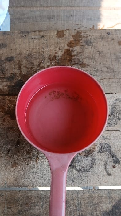 A red plastic ladle, partially filled with a clear liquid containing dark particulate sediment, rests on a weathered wooden surface. The wooden surface comprises multiple planks, displaying visible grain, discoloration, and scattered water droplet stains. In the upper left quadrant, a portion of a light-colored fabric, consistent with a white or light blue garment, is marginally visible. Bright natural light illuminates the scene from the upper right. The observed location is in the city of Bandasafa, Indonesia. No direct human presence or interaction with the ladle is fully discernible.