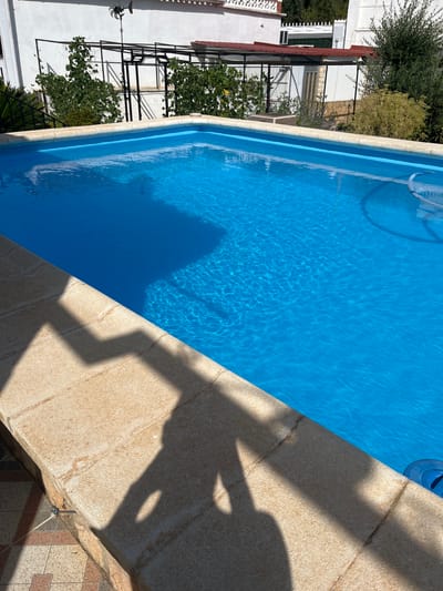 The image shows a close-up, high-angle view of a bright blue swimming pool with rippling water. The pool has a tan-colored coping stone edge. Sunlight casts distinct shadows across the coping stone, indicating it is daytime and likely sunny. To the left of the pool, partially visible, is a white building with some greenery and a black metal structure, possibly a pergola or trellis, with plants growing on it. The scene appears to be an outdoor residential setting. The image is oriented vertically.