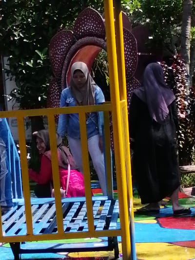 The image shows an outdoor scene, likely in a park or amusement area, with three individuals visible. In the foreground, a bright yellow metal railing runs horizontally and vertically, partially obscuring the view. Behind the railing, on a blue and red painted surface, a young person wearing a pink hijab and a bright pink garment is sitting. To their left, another person, also wearing a hijab and a light blue denim jacket over white pants, stands. Further to the right, a third person, wearing a dark outfit and a purple hijab, stands facing away from the camera. In the background, a large, stylized representation of a Rafflesia flower is visible, along with lush green foliage and a palm tree. The ground is painted with various bright colors, suggesting it may be a play area or a themed attraction. This scene is located in the city of Tegalwangi, Indonesia.