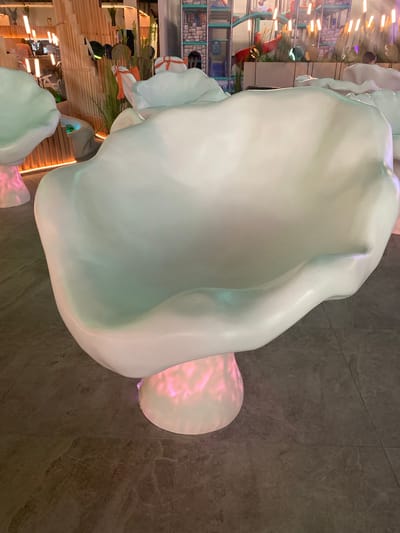 This is a photograph of a large, white, abstract sculpture, possibly a chair or seating area, with soft, diffused lighting that casts a pinkish glow on a portion of it. The sculpture is organic in shape, with smooth, flowing curves and a slightly dimpled surface. It appears to be made of a solid, molded material.
In the background, there are elements suggesting an indoor entertainment or play area. These include what looks like a climbing structure with colorful netting and a slide. There are also wooden slatted walls and overhead lighting fixtures with a warm, yellow glow, reminiscent of strip lights. Some areas of the background show glimpses of what could be decorative elements like artificial plants or foliage, and possibly some artwork or displays.
The overall setting appears to be a modern, brightly lit indoor space, likely a children's play center, a themed cafe, or a public art installation. The image is oriented vertically, making it a portrait-style photograph. The lighting creates a slightly surreal or dreamlike atmosphere. There is no discernible text in the image. The focus is on the sculptural element, which dominates the foreground.