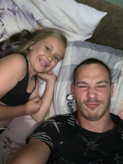 In a dimly lit room, two individuals lie on a bed covered with floral and striped bedding. On the left, a young girl with blonde hair smiles at the camera, resting her arm on a pillow. She wears a black tank top. To the right, a man with short brown hair and stubble squints into the camera, a slight smile on his face. He is wearing a black t-shirt with a graphic print. The scene appears to be a casual, indoor setting, likely a bedroom. The location is Astoni, Latvia.