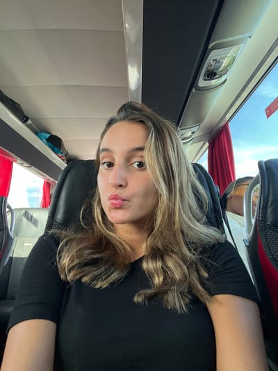 The image depicts the interior of a passenger bus or coach during daylight hours. A female subject occupies the central foreground, seated in a black upholstered seat. She has medium-brown skin, dark eyes, and long, wavy brown hair with blonde highlights. She is wearing a black ribbed short-sleeved top. Her lips are pursed in a pouting expression, and she is looking directly at the camera.
To the subject's right and slightly behind her, a portion of a second individual is visible, seated in an adjacent seat. Only the top of their head, an ear, and a segment of their face are discernible, showing they are wearing glasses and a light-colored top. This individual is oriented away from the camera, seemingly looking towards the bus window or forward.
The bus interior features overhead luggage racks running along the length of the ceiling, with individual passenger lights and air vents. Red curtains are pulled back from the windows on the right side of the bus, revealing a clear blue sky and a suggestion of outdoor foliage. A red informational sticker is affixed to the window frame near the top right corner. A dark-colored bag is visible on the overhead rack on the left side of the bus. The general environment is brightly lit by natural light. The specific location within Cantanhede, Portugal, is not discernible from the visual information provided in the image.