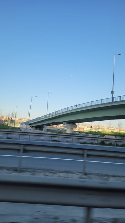The image shows an elevated highway in Beylikdüzü, Turkey. The sky is clear blue. The highway structure is green and gray, with a metal railing. There are street lights alongside the road. Below the elevated highway, there is a road with white lane markings. There are trees and buildings visible in the distance. The immediate foreground contains blurred gray guardrails.
