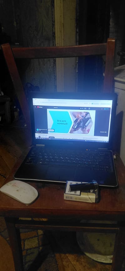 The image depicts a laptop open and displaying a YouTube video, situated on a wooden chair in a dimly lit room. The laptop screen shows a video with Ukrainian text that translates to "Everything for collections." A white computer mouse rests to the left of the laptop, and a pack of cigarettes and a lighter are placed in front of the laptop. The background reveals a wall with patterned wallpaper and a wooden floor. The setting appears to be a personal living space. There are no individuals visible in the image. The overall scene suggests a casual or personal use of the laptop, possibly for research or leisure related to collecting. The location is identified as Ketrysanivka, Ukraine.