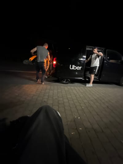 A black Uber vehicle is parked on a cobblestone surface at night. A man wearing a white t-shirt and black shorts stands near the open passenger-side door. Another man wearing a gray shirt and jeans stands near the rear of the vehicle next to a scooter. Several cigarette butts are visible on the cobblestone. A person wearing dark pants and shoes is in the foreground of the image.