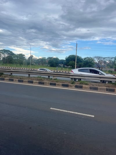 The image depicts a roadside scene located in Kahawa Sukari ward, Kenya, under a sky with significant cloud cover and intermittent patches of blue. In the immediate foreground, a dark, damp asphalt road surface is visible, marked by a single white broken line. A multi-tiered metal guardrail functions as a median barrier. Beyond this barrier, on a raised, unpaved median strip, a light-colored four-door sedan, consistent with a Honda Insight, is positioned on the right side of the frame, partially obscured by the guardrail. An indistinct occupant is visible within the driver's seat. Further to the left on the same median, a white hatchback is stationary, angled slightly towards the right. One individual is observed walking rightward on the unpaved median behind the hatchback. The background consists of a dense line of green trees and foliage, with utility poles (including a street light pole) and overhead power lines intersecting the sky. Faint outlines of distant structures are discernible through the trees.