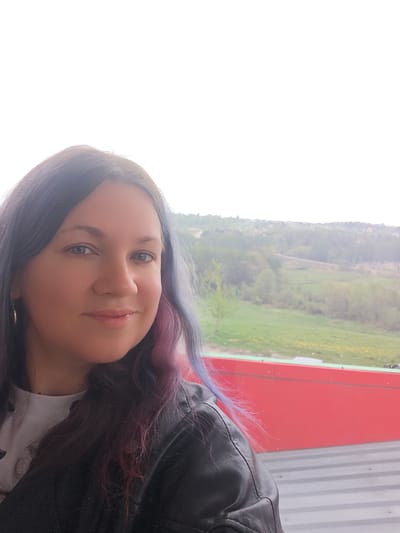 A woman poses for a selfie outdoors in the city of Ильинское, Russia. She is wearing a black leather jacket over a white t-shirt. Her hair is dark with streaks of blue and pink. Behind her is a landscape with a green field, trees, and some buildings in the distance. A bright red structure is visible at the bottom right of the frame. The sky is overcast and white.