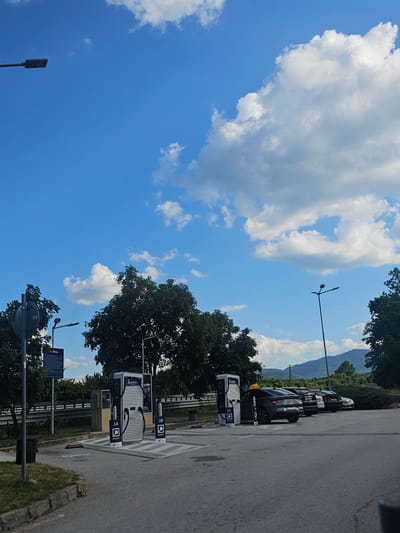 The image shows an outdoor scene, likely a parking lot for electric vehicles, with a clear blue sky and scattered white clouds. In the foreground, there are several electric vehicle charging stations, some labeled with "1A" and "1B" and bearing a green charging symbol. The stations appear to be modern and white with green accents.
Behind the charging stations, a row of parked cars is visible, suggesting they are either charging or waiting to charge. The cars are dark in color, and some have license plates visible, though they are not legible enough to read.
In the background, lush green trees form a dense line, and beyond them, rolling hills or mountains are visible, indicating a scenic or rural location. The time of day appears to be daytime, given the bright sunlight and clear sky. The weather seems pleasant, with a bright sky and sunshine.
There is a lamppost visible in the sky on the left side of the image. Another lamppost with multiple lights is also present in the upper right portion of the image. A blue sign with white text is partially visible on the right side, possibly indicating information or instructions related to the charging station or parking area.
The scene depicts a functional setting for electric vehicle charging, set against a backdrop of natural beauty. No people are visible in the image. The overall mood is calm and orderly.