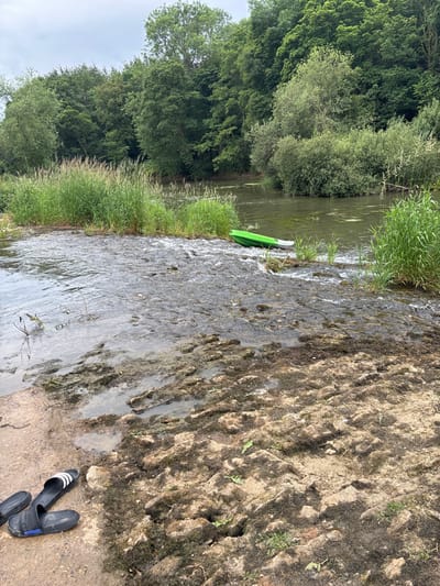 A landscape shows a river flowing beside a grassy bank and a tree-lined shore. A green paddleboard rests at the water's edge. The foreground features a textured, earthy bank leading to the river. Footwear, two black and white slides, lie on the bank's edge. The river's surface is somewhat reflective, showing slight distortion from the flowing water.