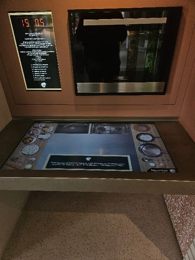 An interior installation in Avignonet-Lauragais, France, features interactive culinary exhibits. The upper left of the scene presents a wall-mounted digital display unit. This unit includes an LED clock displaying "15 05" in red digits, positioned above a black panel with white French text detailing instructions or information, accompanied by a chef's hat icon. To its immediate right, a built-in appliance, likely an oven, with a black glass front and two horizontal brushed metal accents, reflects a blurred, dark-clad individual, along with indistinct green and brown environmental elements.
Directly below these wall-mounted components, an interactive digital tabletop display is integrated into a counter surface. This display renders an overhead view of a virtual kitchen workspace, illustrating various food ingredients (e.g., sliced vegetables, raw meat, grains) arranged on the left periphery, and cooking utensils (e.g., frying pans, spatulas) on the right. A central gray area within the display represents a cooking surface. A textual overlay in white, with French instructions and a chef's hat icon, provides context within the interactive display. The surrounding wall and counter surfaces are a uniform reddish-brown. The lower surface, appearing as a floor or base cabinet, is a mottled gray-brown with dark flecks. A section of patterned wallpaper is partially visible on the far right.