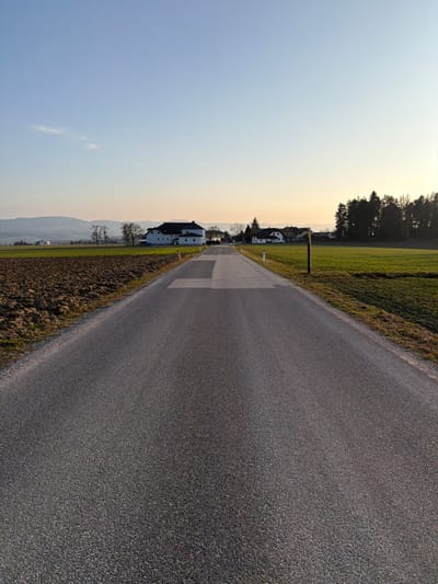 The image depicts a rural road in Ferschnitz, Austria, extending straight from the foreground towards a distant cluster of buildings under a late afternoon or early evening sky.

The foreground is dominated by a dark grey asphalt road, appearing to be two lanes wide, without painted markings. Approximately one-third of the way into the frame, several lighter rectangular patches are visible on the road surface, indicative of repairs or faded paint.

To the left of the road, a narrow strip of green grass borders a large, dark brown, freshly tilled agricultural field. Beyond this field, in the middle distance, stands a prominent two-story building with light-colored walls and a dark roof, accompanied by several other structures and leafless deciduous trees. Distant rolling hills are visible on the horizon.

To the right of the road, a similar narrow strip of green grass separates the asphalt from a vibrant green field, likely pasture or young crops. A brown and white reflective marker post stands vertically on the right shoulder of the road, roughly parallel to the lighter patches on the road surface. Further in the distance, a dense grouping of dark evergreen trees forms a distinct boundary against the horizon.

The sky transitions from a clear light blue directly overhead to a soft yellow and orange glow near the horizon, consistent with sunset or sunrise. A minimal, wispy cloud is discernible in the upper left quadrant. No people, vehicles, or animals are present in the scene.