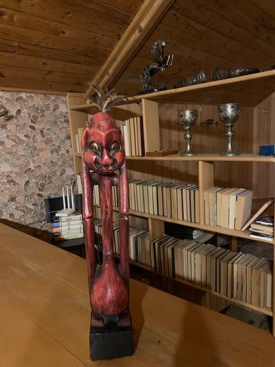 The image depicts the interior of a room with a wooden sloped ceiling. In the foreground, a tall, red, wooden sculpture of a figure with long limbs and a stylized face stands on a wooden table. Behind the sculpture, a bookshelf is filled with numerous books. The shelves also display decorative items, including two silver chalices and a bronze sculpture of a horse. To the left of the bookshelf, a stack of books is topped with a white wireless router. The wall behind the books and router is covered in a patterned wallpaper. The scene is dimly lit, suggesting it may be evening or nighttime. No people are visible in the image. The location is identified as Mepiskalaki, Georgia.