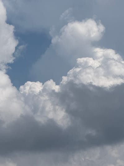 The image shows a cloudy sky. Cumulus clouds, some bright white and others dark gray, fill the frame. A patch of blue sky is visible towards the upper left. The image does not depict any specific location in the city of Đa Lộc, Vietnam, nor does it contain any people, subjects, objects, or actions. It is a natural atmospheric scene.