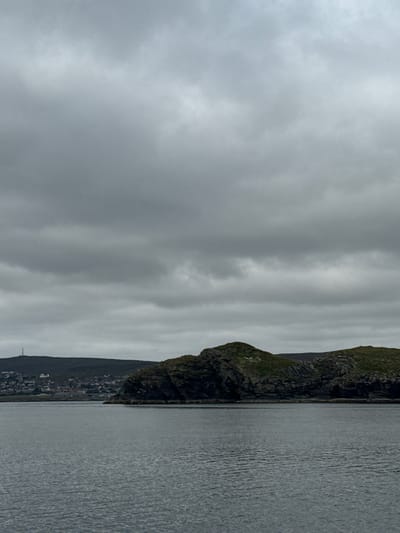 The image shows a body of water in the foreground, with a rocky island or peninsula in the middle ground. On the far left of the image, a coastal town with buildings and a tall antenna is visible on a hillside. The sky is overcast with dark, heavy clouds. The overall scene depicts a coastal landscape under a gloomy sky. The town on the left is identifiable as part of Lerwick, Shetland Islands, United Kingdom. The rocky formation in the middle ground is likely a part of the coastline near Lerwick.