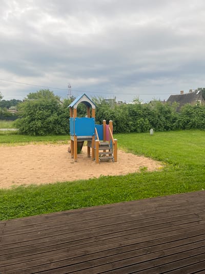 The image depicts an outdoor children's playground with a sandbox and a small play structure. The play structure is made of wood and plastic, featuring a blue roof and blue panels. A set of wooden stairs leads up to a platform. The playground is situated on a patch of sand, bordered by green grass. In the background, there are dense green bushes and trees, some houses with visible rooftops, and a tall communication tower. The sky is overcast. The foreground shows a dark wooden deck. The scene is a typical urban or suburban environment, and it is plausible that this playground is located in the city of Narva, Estonia, given its architectural style and vegetation. No people are visible in the image.