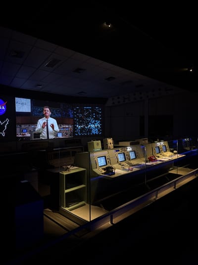 The image depicts a dimly lit, simulated mission control center, specifically located within an exhibition in Canela, Brazil. The scene features a large projection screen on the rear wall displaying a male presenter in a white collared shirt and dark tie, gesturing with both hands, flanked by graphical data, including a NASA logo and complex charts. In the midground and foreground, a series of vintage-style control consoles, light gray in color, are arranged. Each console is equipped with multiple square CRT monitors displaying blue graphical information. Objects on the consoles include a black rotary telephone, a red push-button telephone, and stacks of papers. A clear glass barrier separates the viewer's area from the control consoles. Overhead lighting fixtures are visible on the dark ceiling, casting focused illumination onto the consoles and creating reflections on the glass barrier. The overall environment suggests a museum exhibit designed to recreate a historical space operations setting. There are no live individuals present within the frame beyond the projected image.