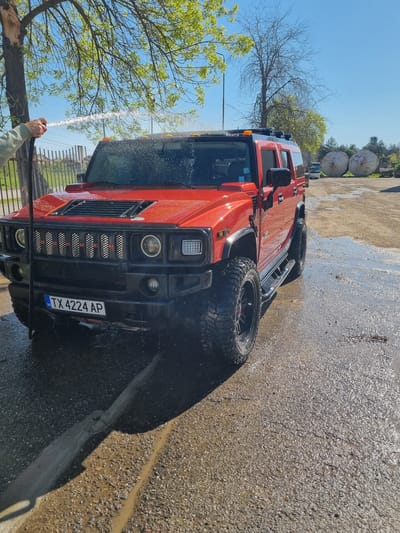 A bright red Hummer H2 SUV is being hand-washed outdoors on a clear, sunny day in Ovcharovo, Bulgaria. The vehicle, positioned prominently in the frame, features large, rugged off-road tires with black rims and a distinctive front grille displaying the word "HUMMER" in individual letters. Square headlights and turn signals are visible on the front. A person's arm, clad in a light-colored sleeve, is visible on the left, holding a hose from which a stream of water is actively spraying onto the vehicle's hood and windshield.
The ground beneath and around the Hummer is a mix of gravel and dirt, appearing extensively wet and muddy with puddles and reflections from the bright sunlight, indicating the ongoing washing activity. A concrete curb or barrier runs along the right side of the wet ground.
In the background, against a clear blue sky, are trees with both bare branches and fresh green leaves, suggesting spring or early summer. Further back and to the right, two large, cylindrical industrial tanks or drums are visible, hinting at an industrial or semi-rural setting. The vehicle's license plate, "BG TX 4224 AP," is clearly legible on the front bumper, confirming its Bulgarian registration. The strong sunlight and clear skies suggest it is daytime, likely morning or early afternoon.