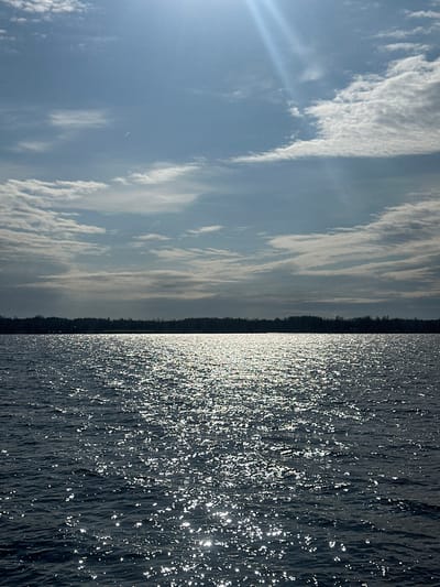 The original image, captured in portrait orientation, depicts a serene natural landscape when viewed rotated 90 degrees clockwise.

The upper portion of the image features a bright, light blue sky adorned with elongated white clouds, suggesting cirrus or altocumulus formations stretching horizontally. Distinct sun rays emanate from the upper-left corner, indicating the sun is positioned just outside the frame, casting strong light. A faint, thin white line, possibly a distant contrail or high-altitude cloud, is subtly visible in the far sky.

A narrow, dark silhouette of a forested landmass forms the horizon line, extending across the mid-ground and separating the expansive sky from the body of water below. This land appears distant and relatively flat.

The lower half of the image is dominated by a dark body of water, likely a lake or river. Its surface is densely textured with countless bright, shimmering reflections of sunlight, creating a dynamic, sparkling effect. This suggests the presence of small ripples or waves under a clear, sunny sky.

The image conveys a clear, sunny daytime scene. No specific activities or individuals are visible, emphasizing the tranquil natural environment. Based on the provided context, the location is Ikšķile, Latvia, suggesting this is a view of the Daugava River or a nearby water body. There is no visible text in the image.
