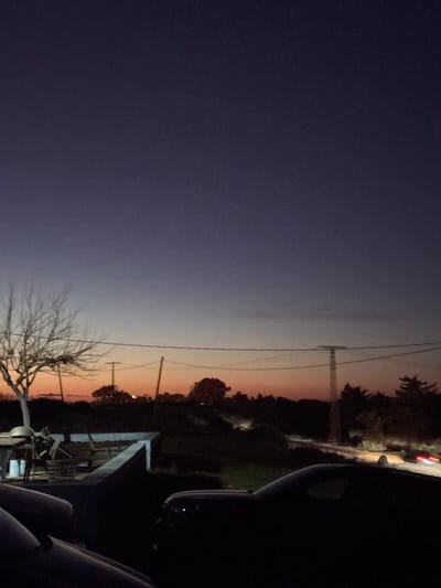 A twilight scene reveals a gradient sky transitioning from dark blue to orange near the horizon. Power lines run across the sky, and trees and structures are silhouetted against the fading light. The view is partially obstructed by the interior of a vehicle, with a dark dashboard and seats visible in the foreground, and the red tail lights of a car in front. The scene conveys a sense of stillness and the end of the day.